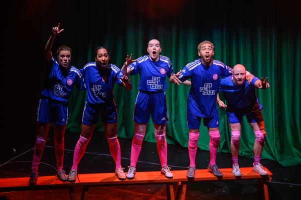 Five people wearing blue football kits stand on a bench looking shocked, on a stage